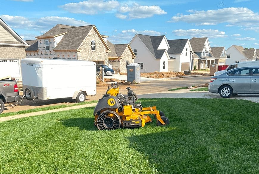 A commercial lawn mower parked on the grass by a suburban street with houses under construction in the background.