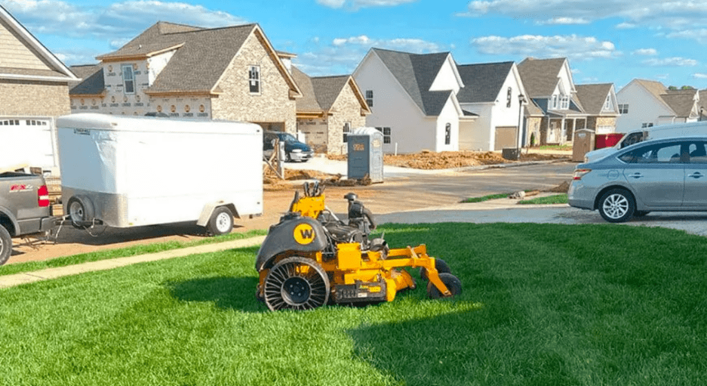 A yellow stump grinder parked on the grass in a residential area with new homes under construction and vehicles around.