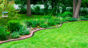 A neatly manicured garden with a curved flower bed and a bird feeder. Tall trees and a white fence are in the background.